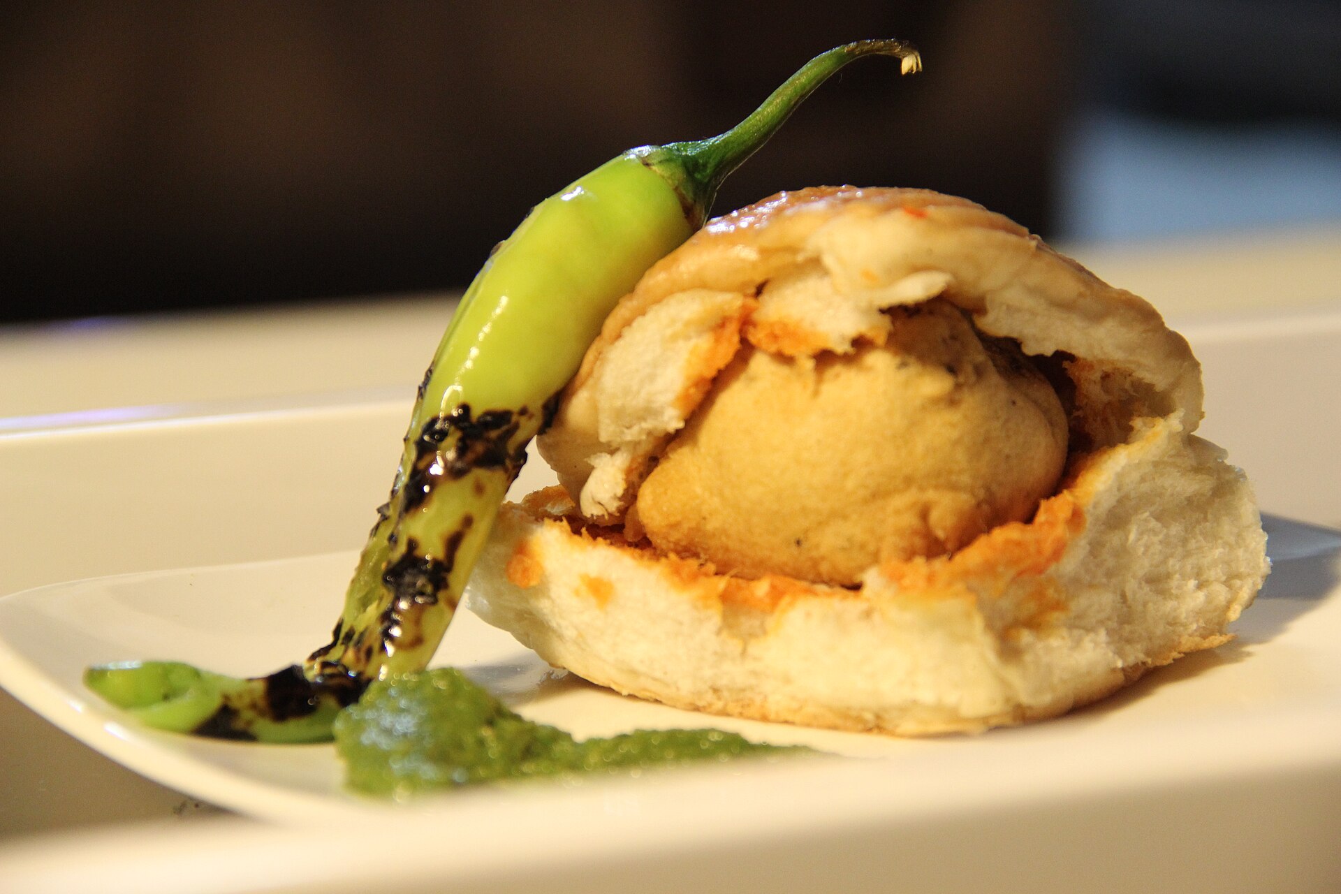 Maharashtrian Street Food | The India A Vada Pav rests on a white plate with green chili and chutney, showcasing delicious Indian street food.