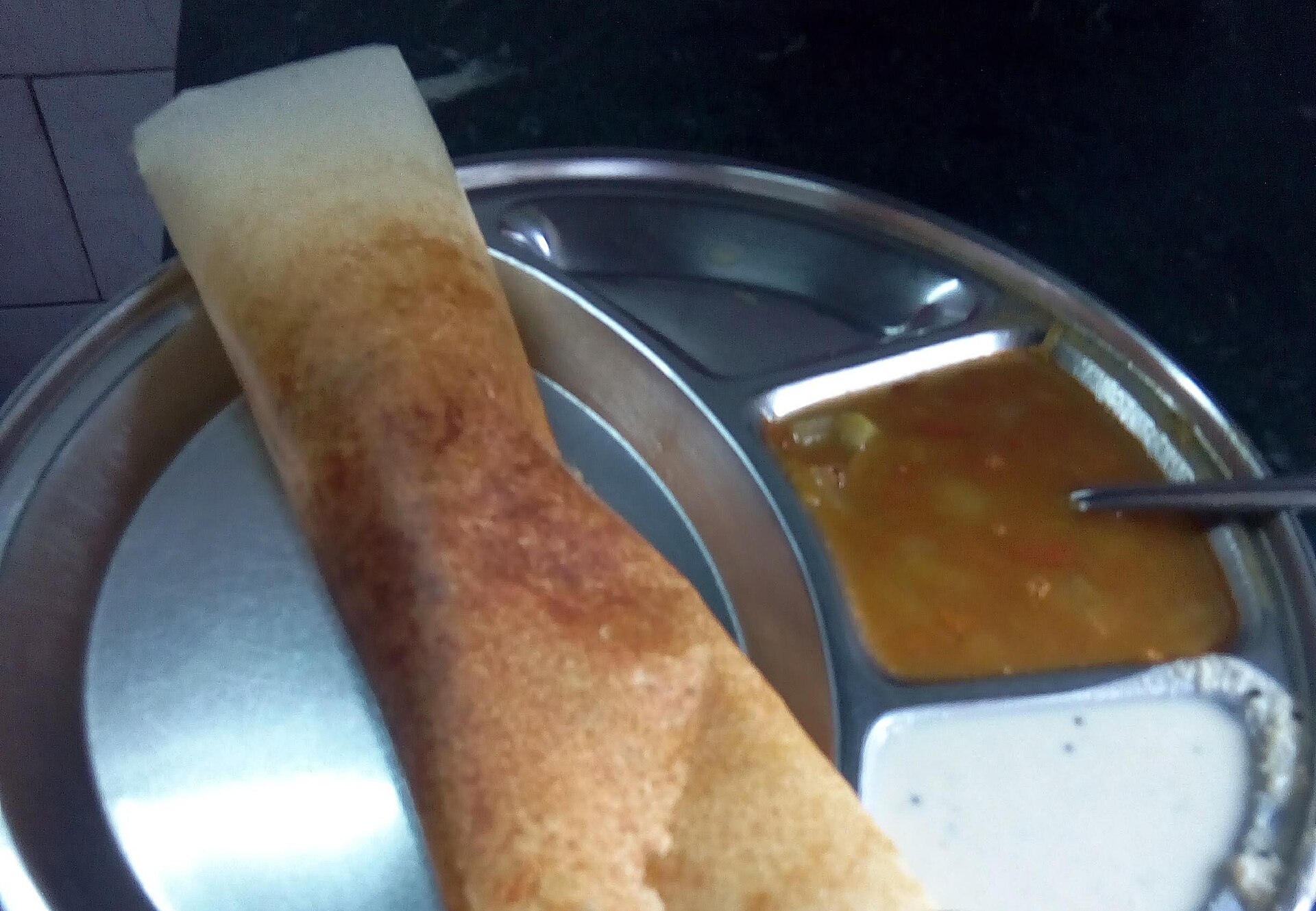 A stainless steel plate holds a crispy masala dosa served with sambar and chutney, showcasing delicious indian food.